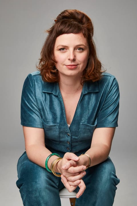 Portrait of Alice Stevens smiling, wearing a blue jumpsuit, against a grey background