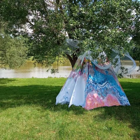 La tente peinte installée sur l’herbe sous un grand arbre, au bord de l’eau