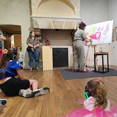 In a museum room, Alice Stevens paints at an easel in front of children sitting on the floor wearing headphones